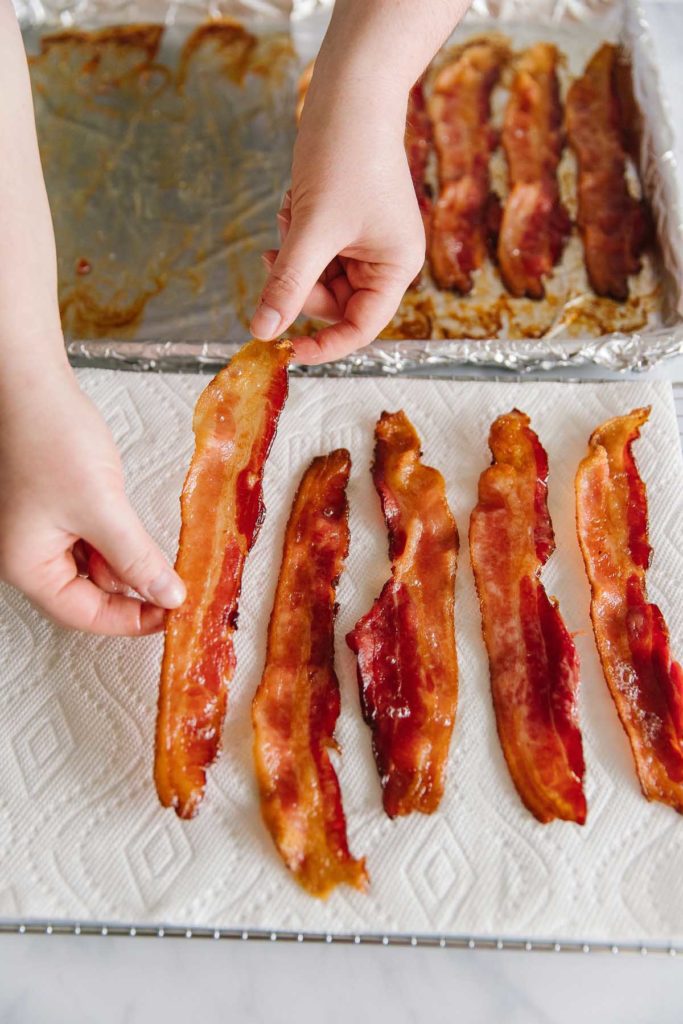 Strips of bacon on a foil-wrapped pan in the back and hands putting strips of bacon on a paper towel.