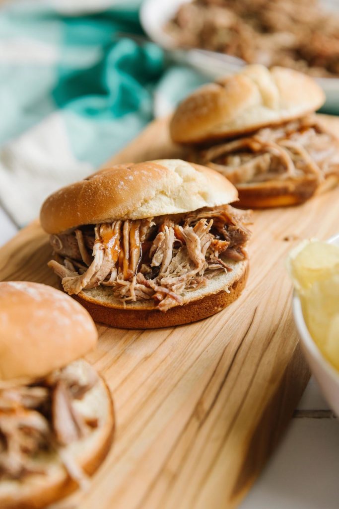There is a wooden plank holding three pulled pork sliders crock pot recipes. In the background you can see a green and white towel and a bowl of chips in the bottom.