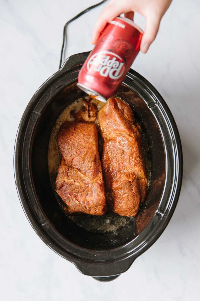 In a black crock pot there are two pieces of raw pork with a hand pouring a can of Dr. Pepper into the crock pot.