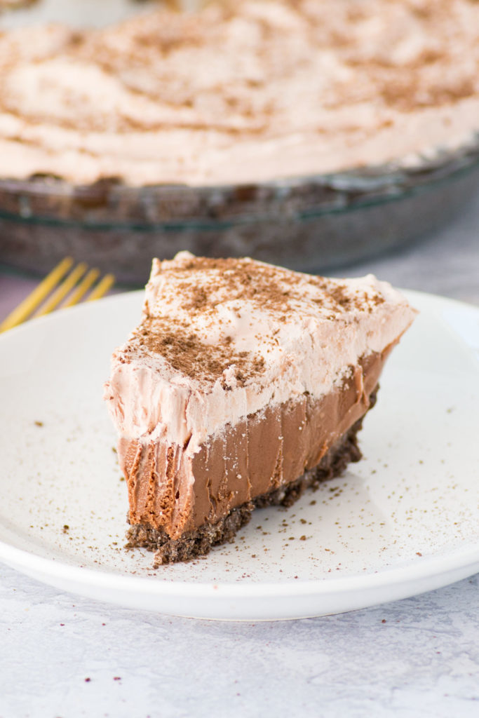 There is one slice of ice cream pie with chocolate crust on a white plate with a gold fork on the side. The rest of the pie is in the back.