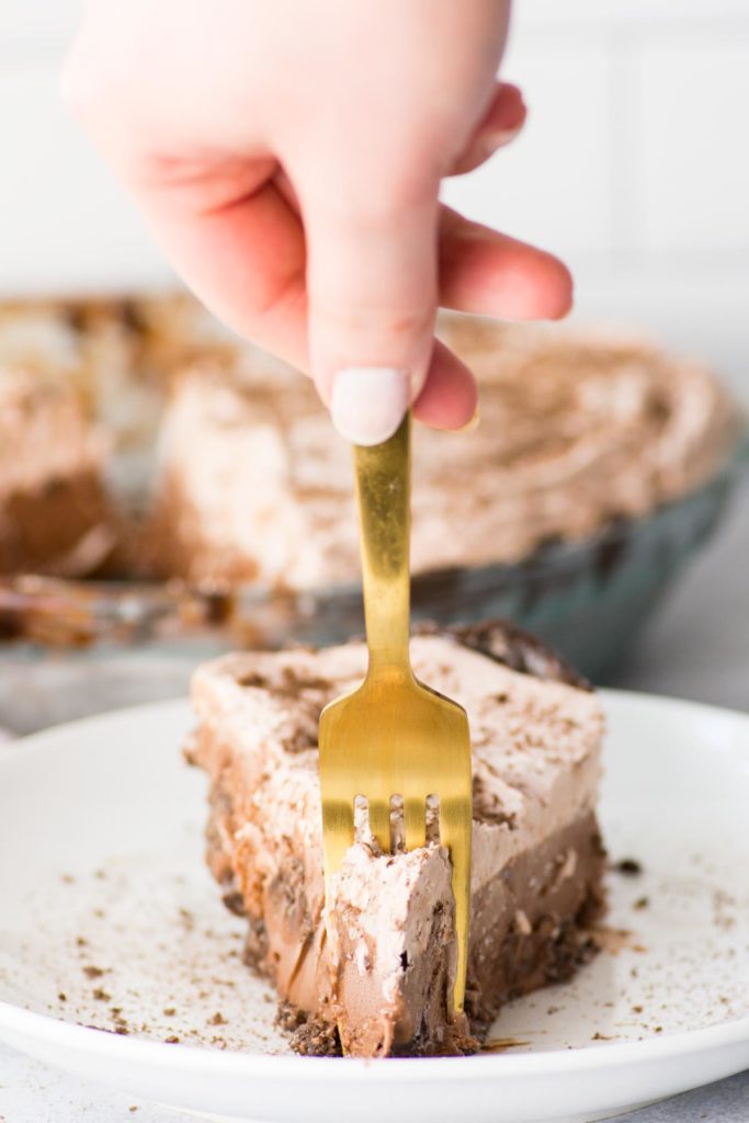 There is one slice of ice cream pie with chocolate crust on a white plate with a hand holding a gold fork piercing the  top of the pie. 