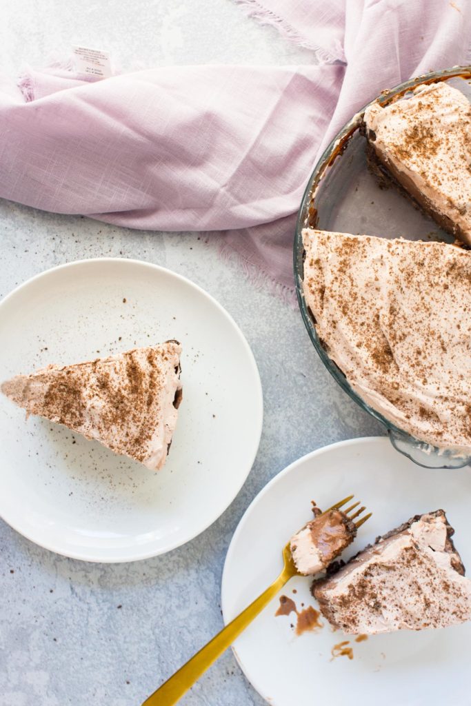 There is one slice of ice cream pie on a white plate with a gold fork. There is one slice of ice cream pie on a white plate on the left side of the picture. The rest of the pie is on the back right side with a pink towel next to it.