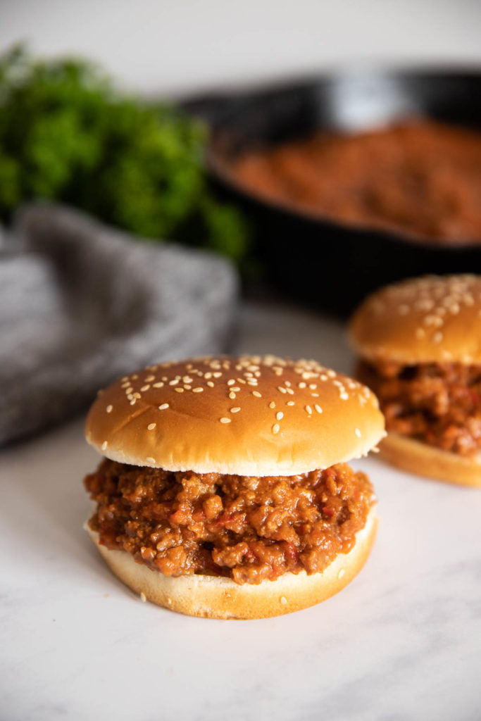 A sloppy joe on a sesame bun with another one behind it with a cast iron pan and green herbs in the back.