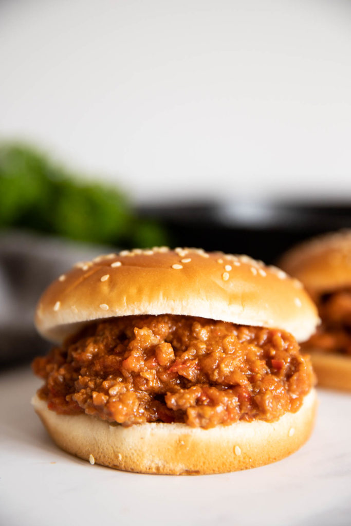 A sloppy joe on a sesame bun with a blurred background with another one behind and green herbs in the back.