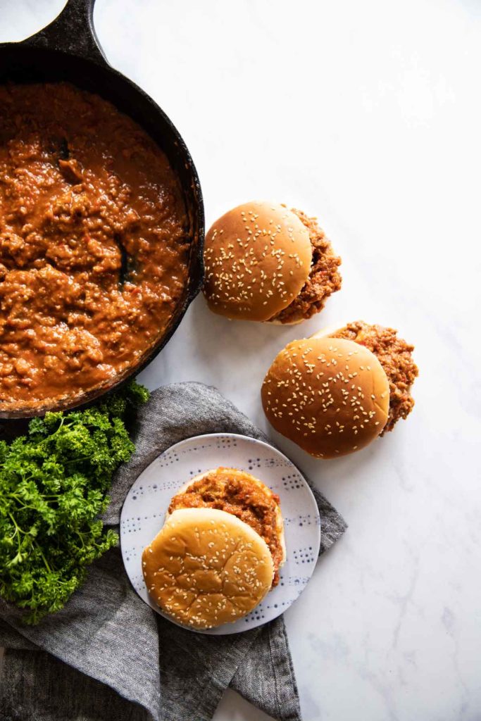 Two sloppy joes on sesame buns are on the counter while one sloppy joe is on a plate on top of a gray napkin with a cast iron pan filled with the mixture and green herbs.