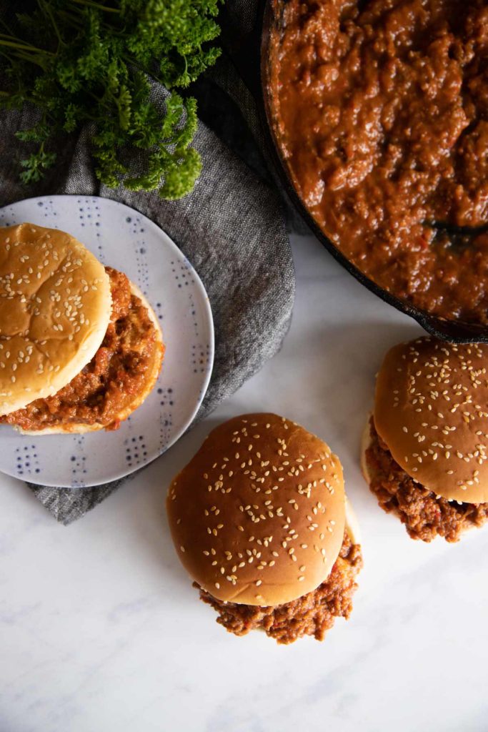 Two sloppy joes on sesame buns are on the counter while one sloppy joe is on a plate on top of a gray napkin with a cast iron pan filled with the mixture and green herbs next to it.