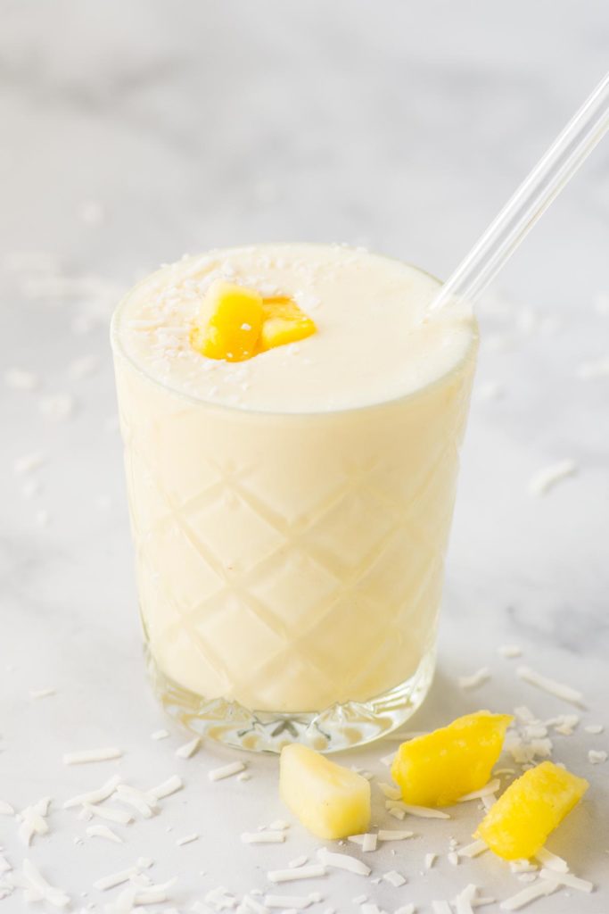 A yellow smoothie in a clear glass with a slices of pineapple and coconut on the side.