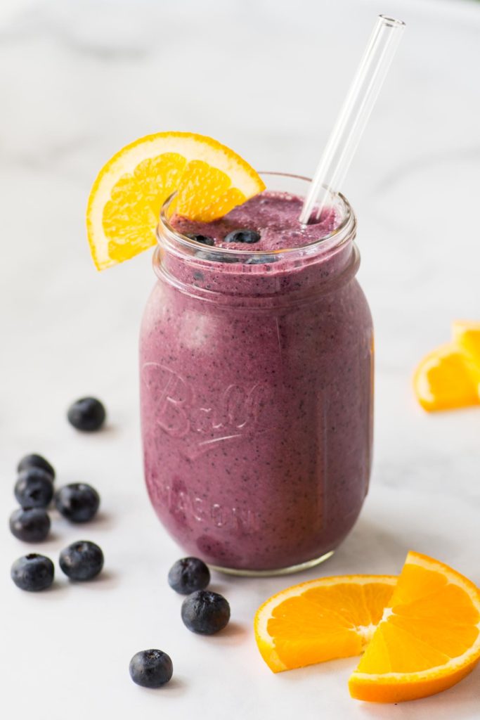 A purple smoothie in a clear glass with a straw and blueberries and orange slices on the side.
