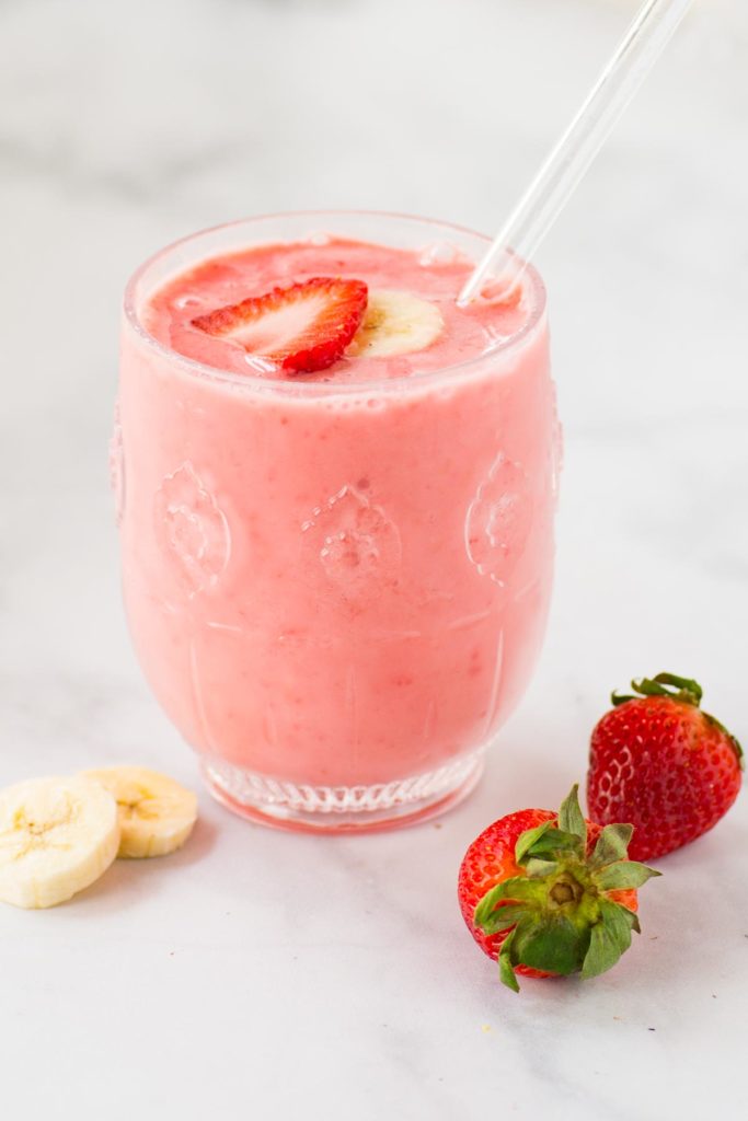 A pink smoothie in a clear glass with a straw with strawberries and banana slices on the side.
