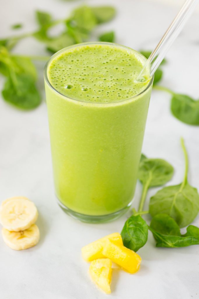 A green smoothie in a clear glass with a straw with spinach, banana slices and pineapple slices on the side.