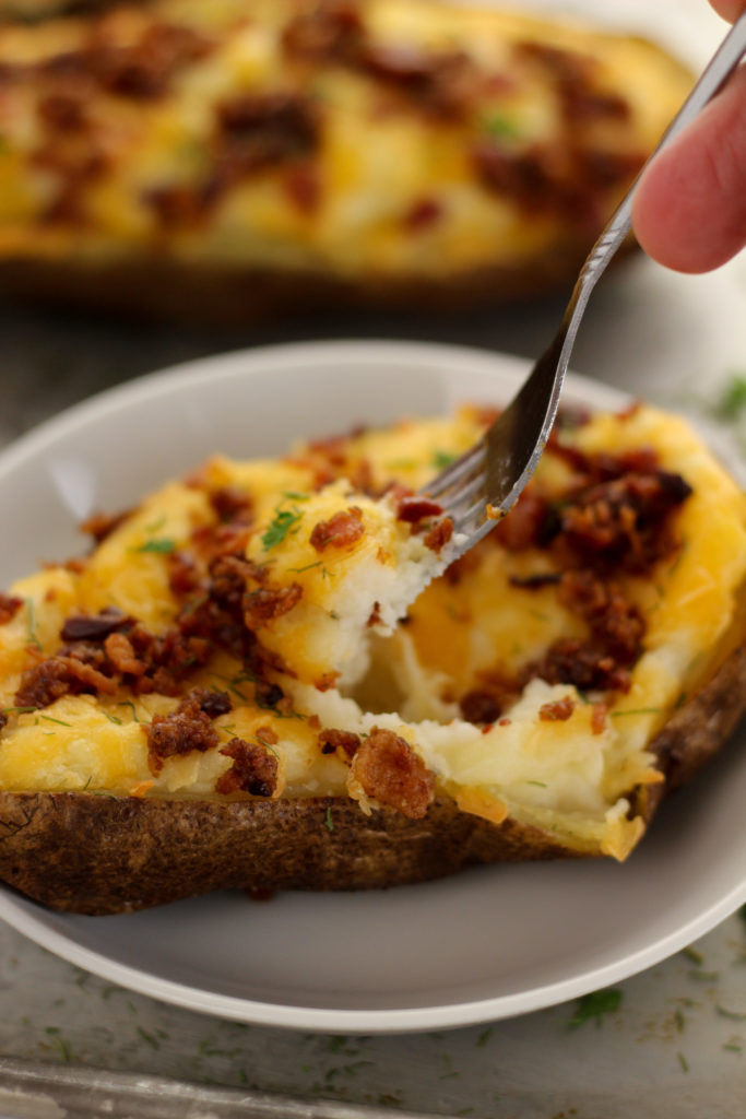 One baked potato topped with bacon and cheese on a white plate with a fork and a hand holding it.  There is another one in the background.