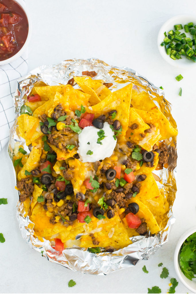 Nachos in a foil packet with a blue and white checked towel with small white bowls full of salsa, jalapenos and cilantro.