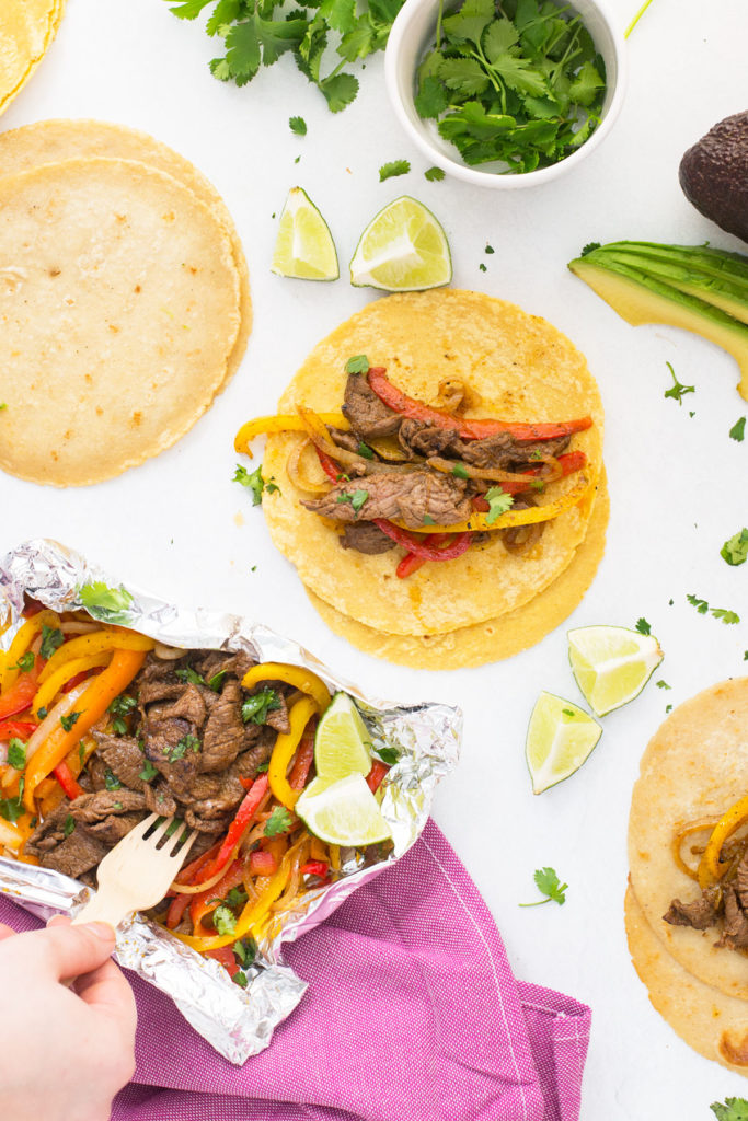 Three stacks of flour tortillas - one plain and two with steak, peppers and onions on it. There is a foil packet with fajita toppings under a pink towel with a hand holding a fork in it and cut up limes around them.