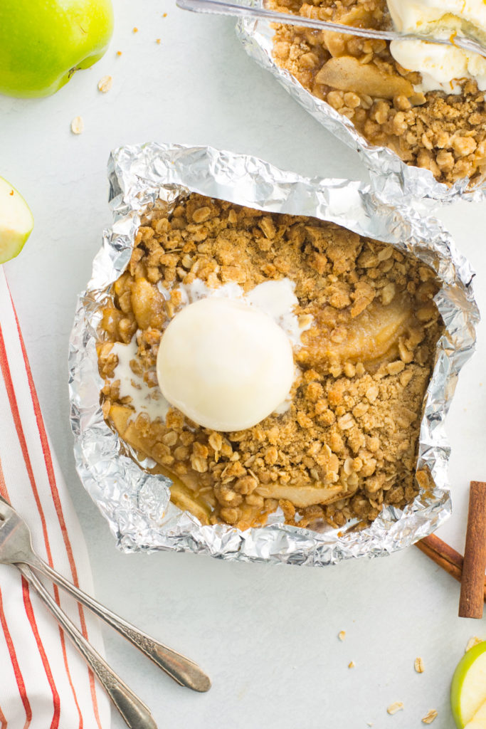 A foil packet filled with apple crisp with a scoop of ice cream and another packet of apple crisp behind it with a spoon in it. There is a green apple and two forks and cinnamon sticks around it.