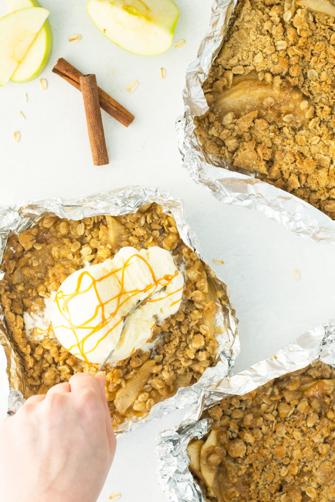 A foil packet filled with warm apples and a cinnamon crumb topping with a scoop of ice cream and a caramel drizzle on it with a fork digging into it with 2 other foil packets of warm apples and a cinnamon crumb topping above and below it.  There is a green apple and cinnamon sticks laying beside it too.