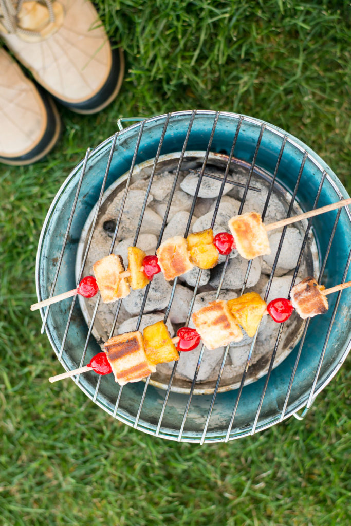A small barbecue with 2 dessert kabobs laying on the grate with the green grass and a pair of boots on the side.