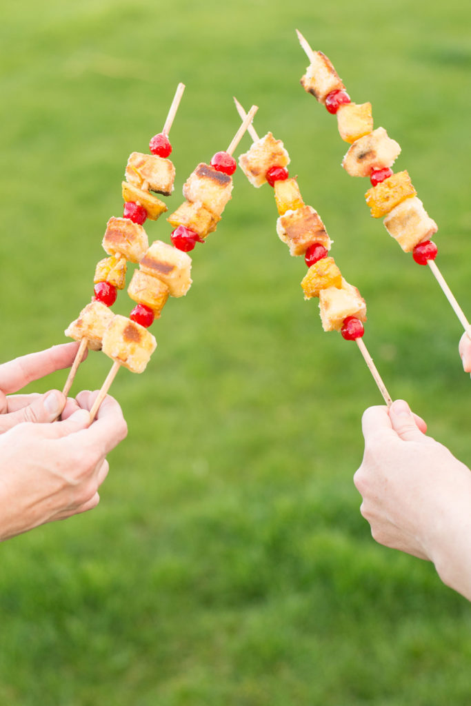 Hands holding pineapple campfire dessert kabobs with green grass in the backyard.