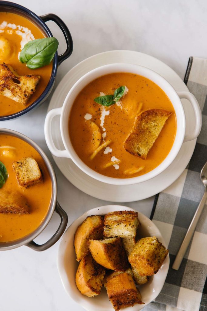 Creamy tomato basil soup in three bowls. Inside bowls of soup are basil, croutons, ravioli and a dripping of fresh cream to garnish.