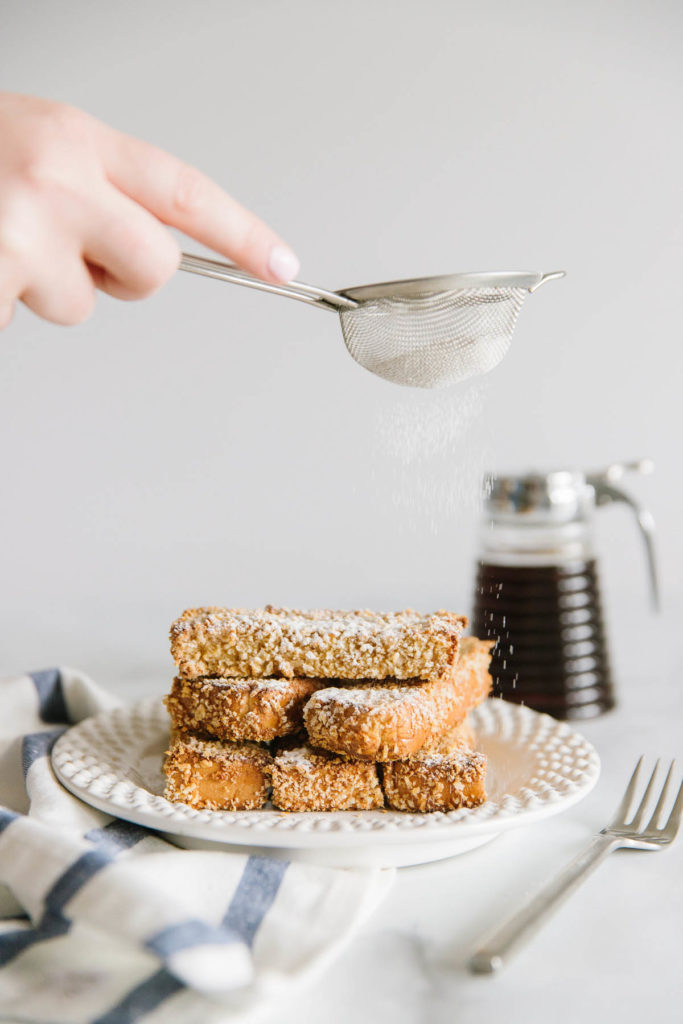 A white plate with 5 French toast sticks stacked on top of each other with a hand sprinkling powdered sugar through a strainer with syrup and a white and blue towel on the side.