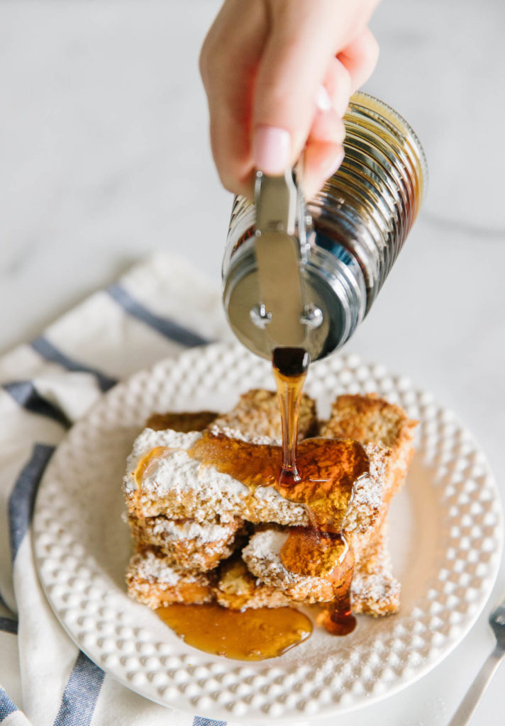A white plate with 5 French toast sticks stacked on top of each other with a powdered sugar and a hand pouring syrup on top with a white and blue towel underneath.