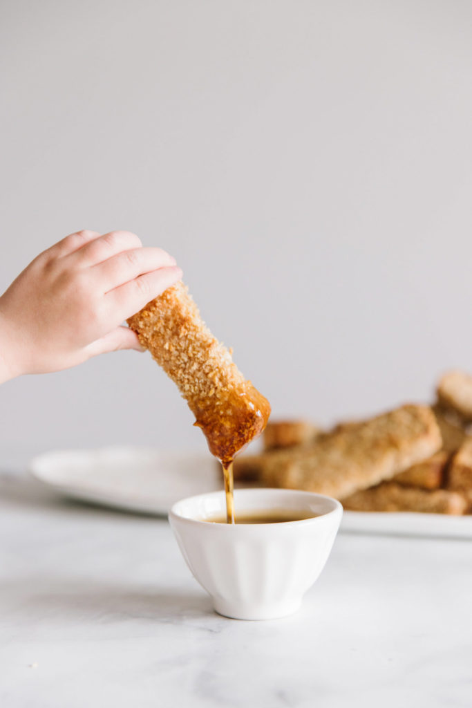 A child's hand dipping a french toast stick into a white bowl of syrup. In the back is a plate full of french toast.