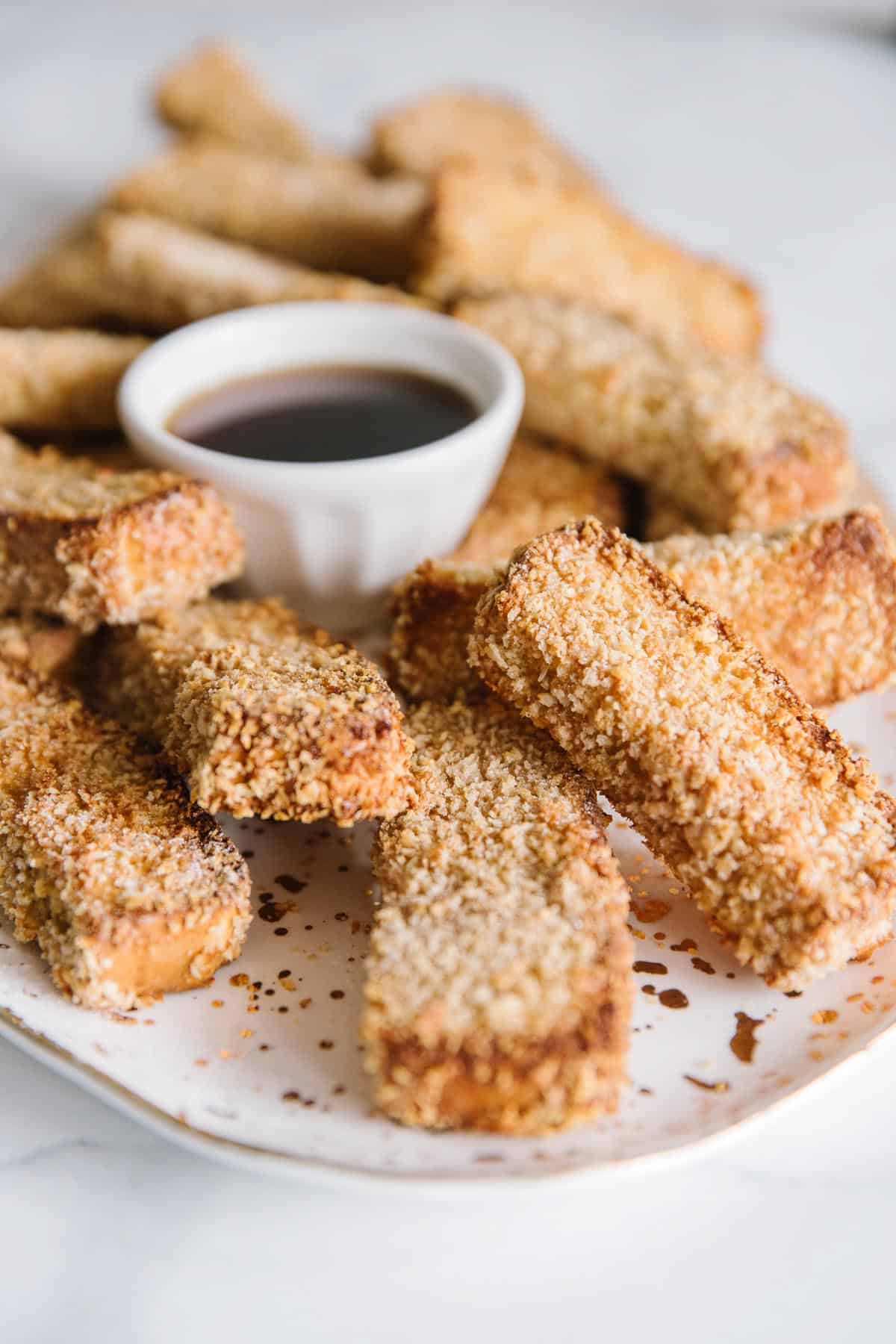 A white plate full of french toast sticks with a white bowl of syrup