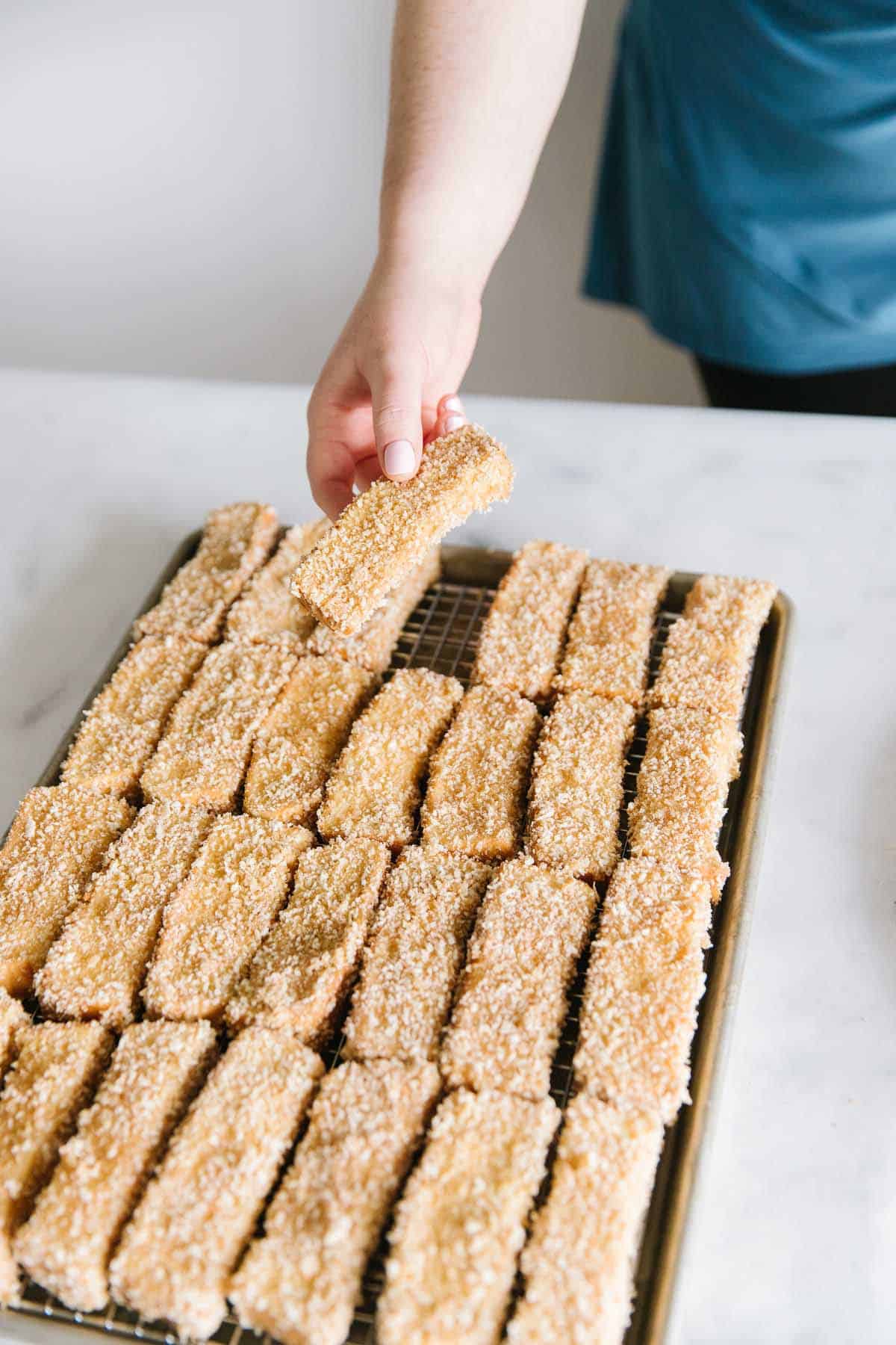 A cookie sheet with french toast sticks laying on the wire rack with one hand holding one stick above.