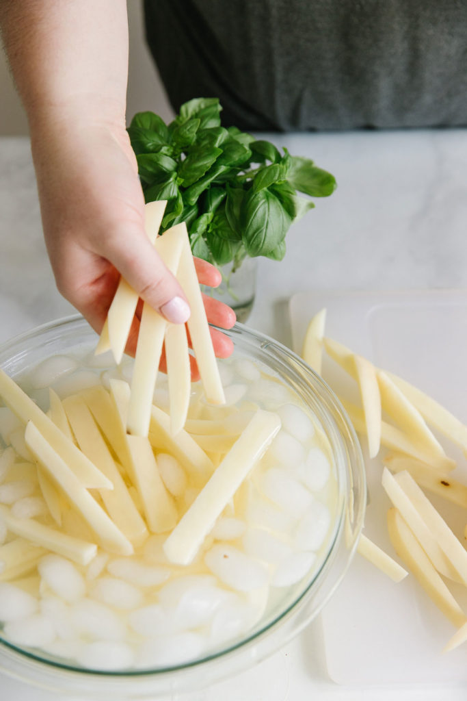 Cut potatoes in a clear glass bowl with water and ice cubes with a hand holding potato slices and basil on the side.