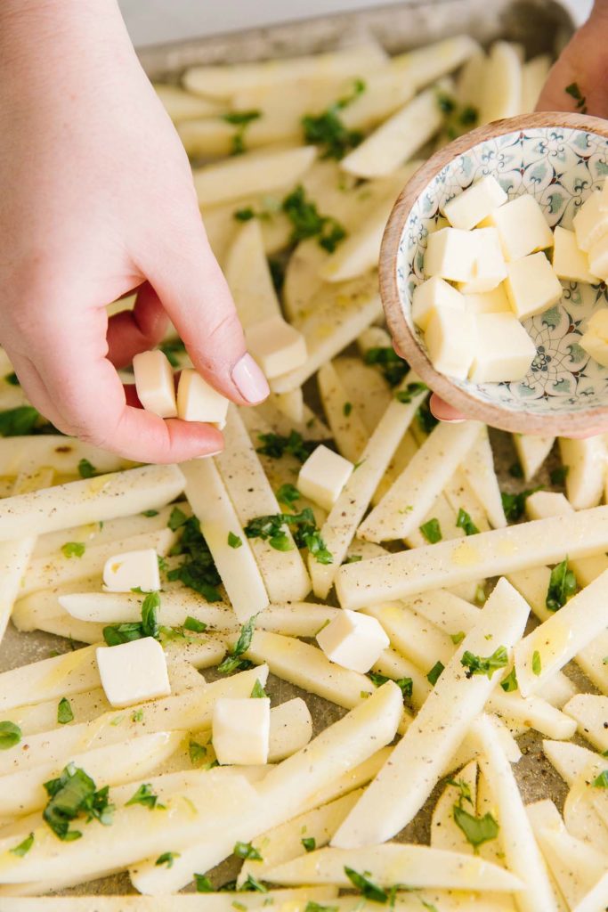 Cut potatoes sprinkled with basil, butter cubes and seasonings on a cookie sheet with two hands holding butter cubes and a bowl of butter cubes.