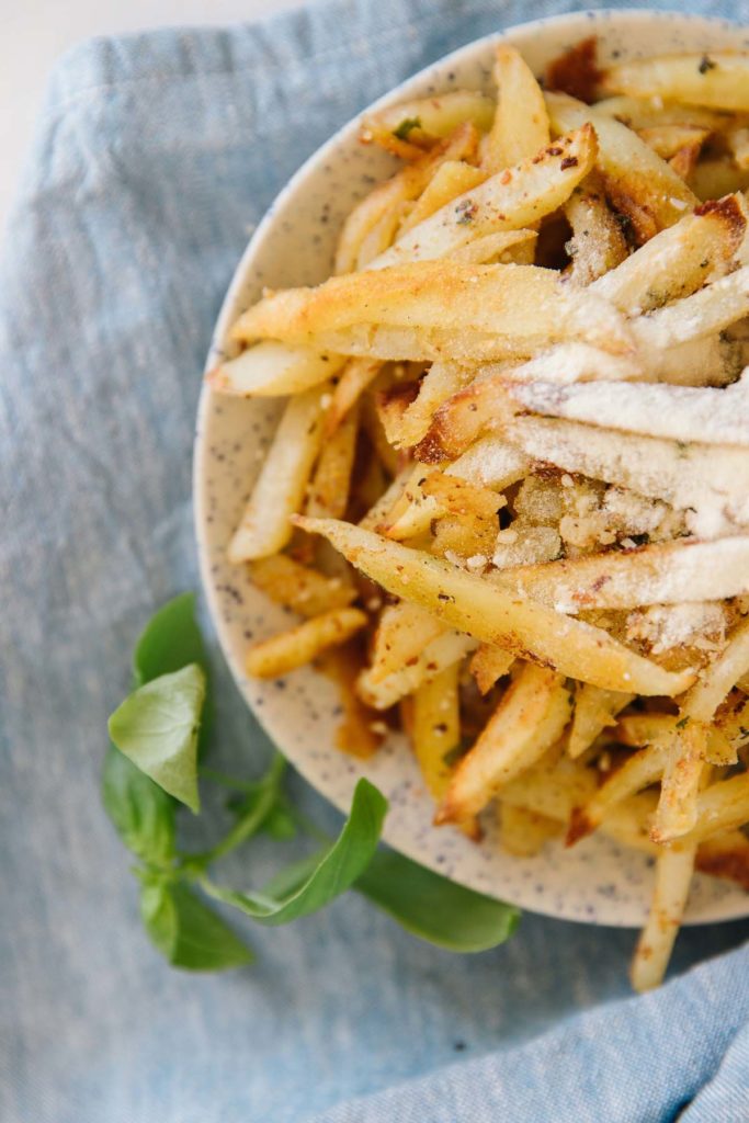 A plate full of french fries with Parmesan cheese sprinkled on it with basil on the side on top of a blue tablecloth.