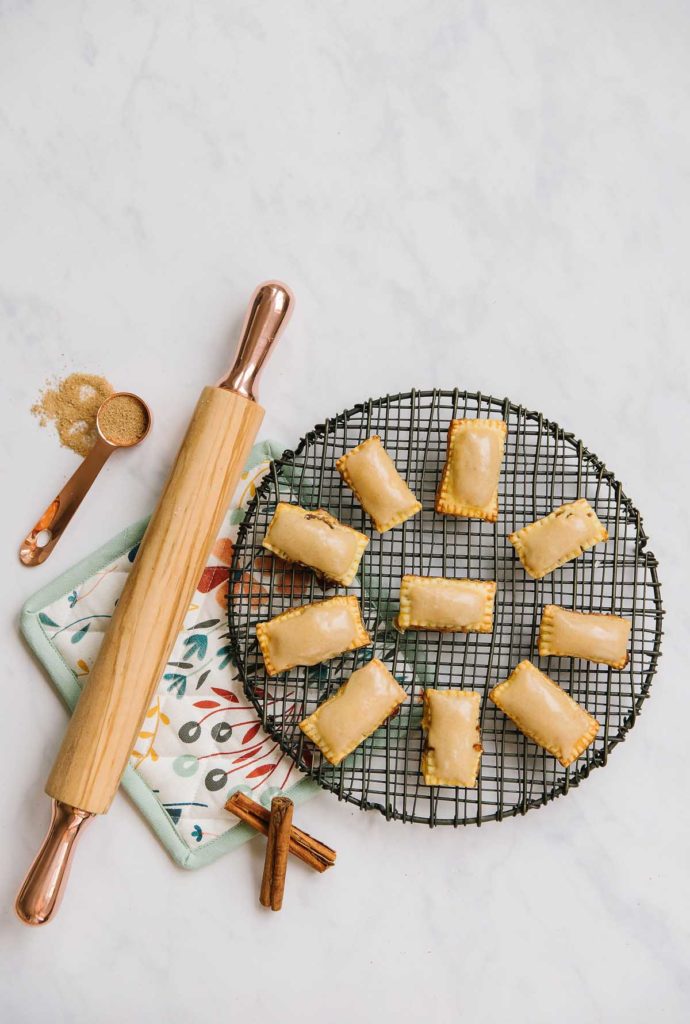 A wire plate with homemade pop tarts with brown sugar icing on them next to a wooden rolling pin, two cinnamon sticks, a measuring cup with brown sugar in it and a flowered hot pad.