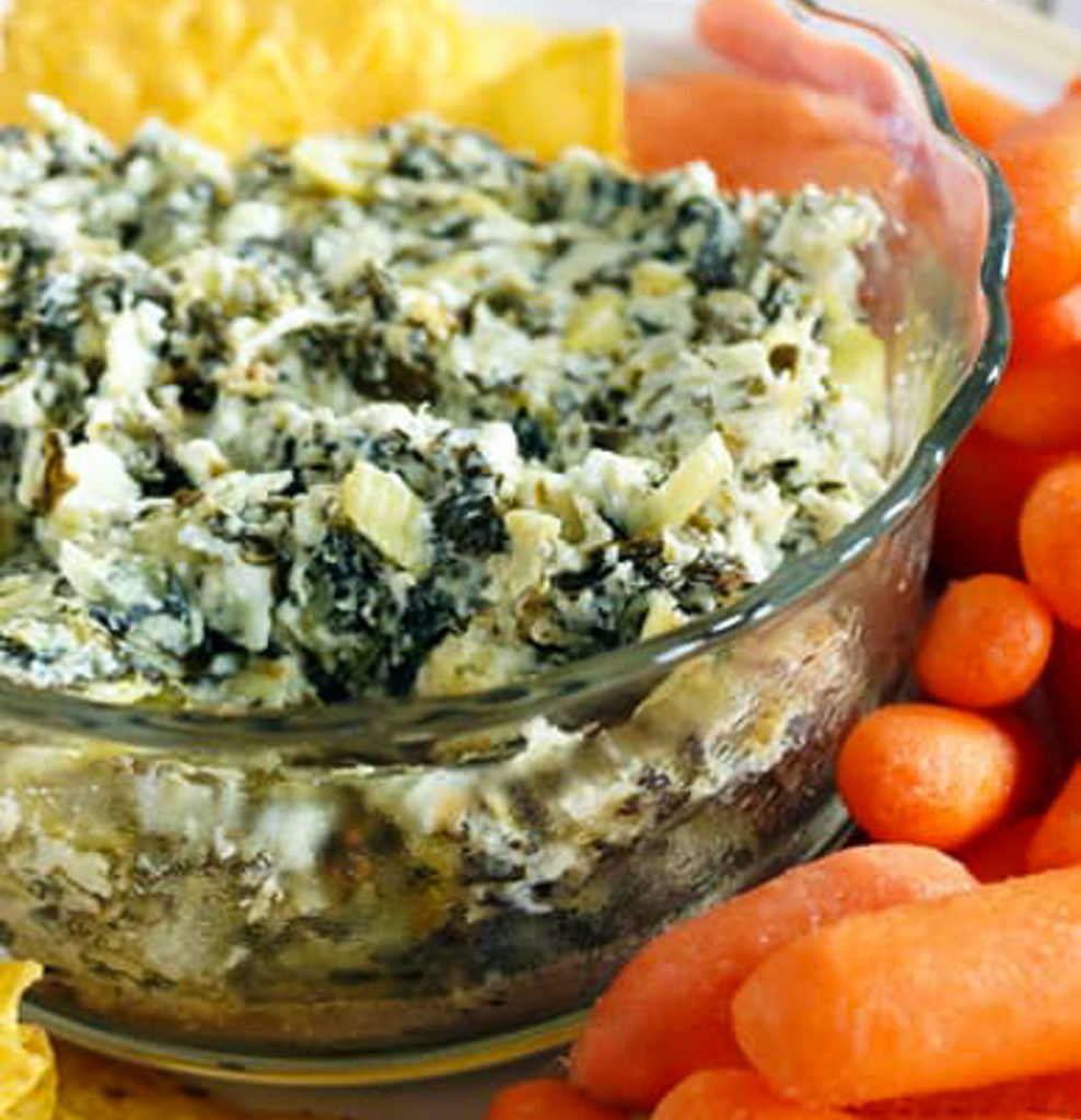 clear glass bowl of artichoke dip and baby carrots on outside of bowl. 