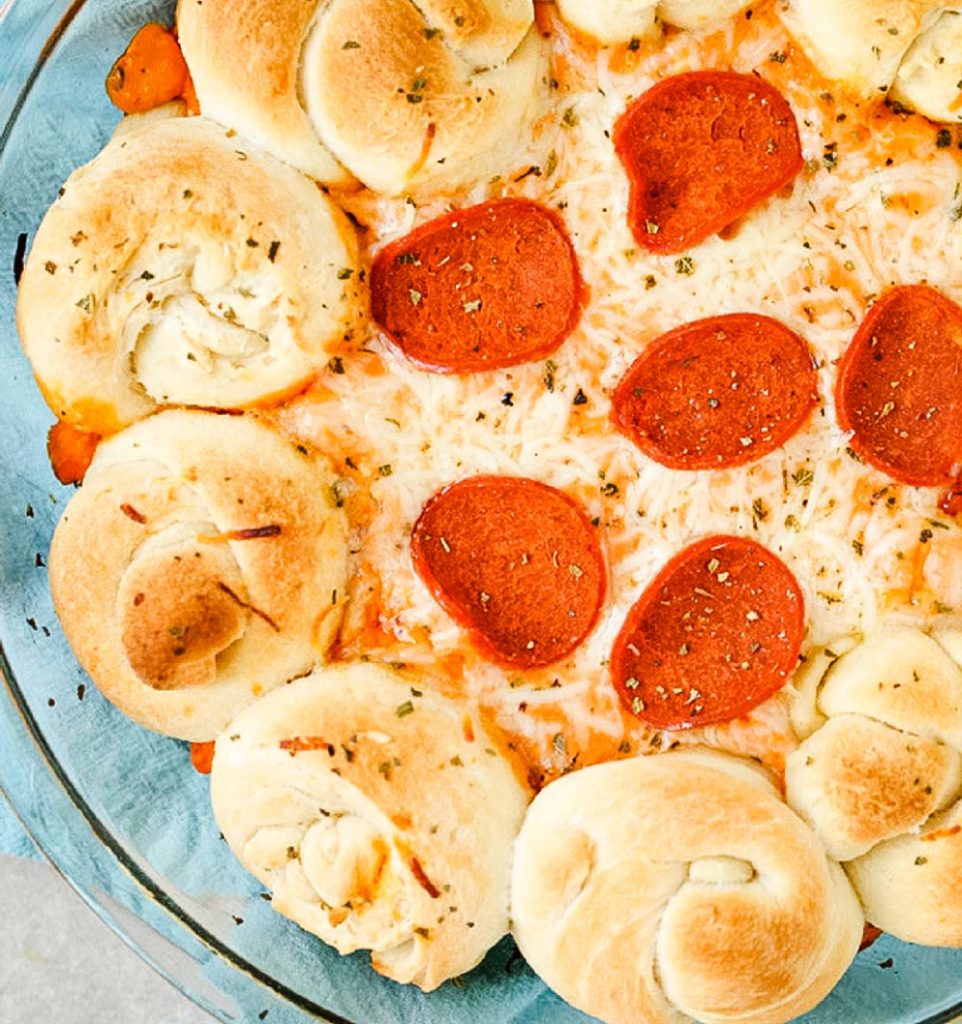 dip with knotted rolls on edges and pepperoni on top in a clear pan. 