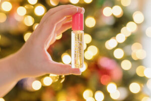 Person holding a container of merry and bright holiday roll-on perfume in from of Christmas lights.