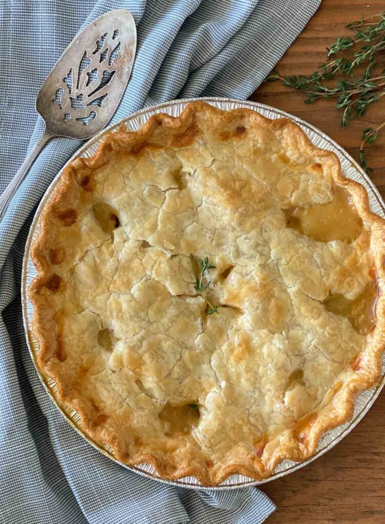 A whole chicken pot pie is on a wooden table with a sprig of herbs, a blue tablecloth and a serving utensil.
