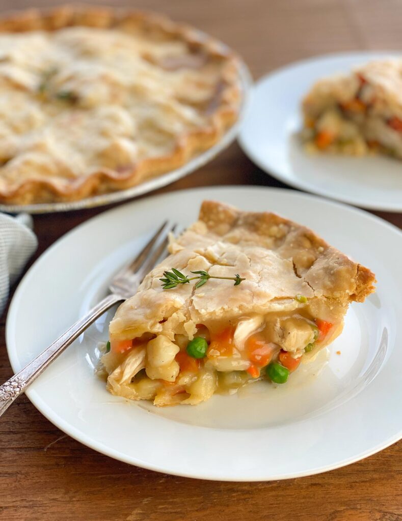 Chicken Pot pie on a white plate with a fork with a whole pie in the background and another slice behind it.
