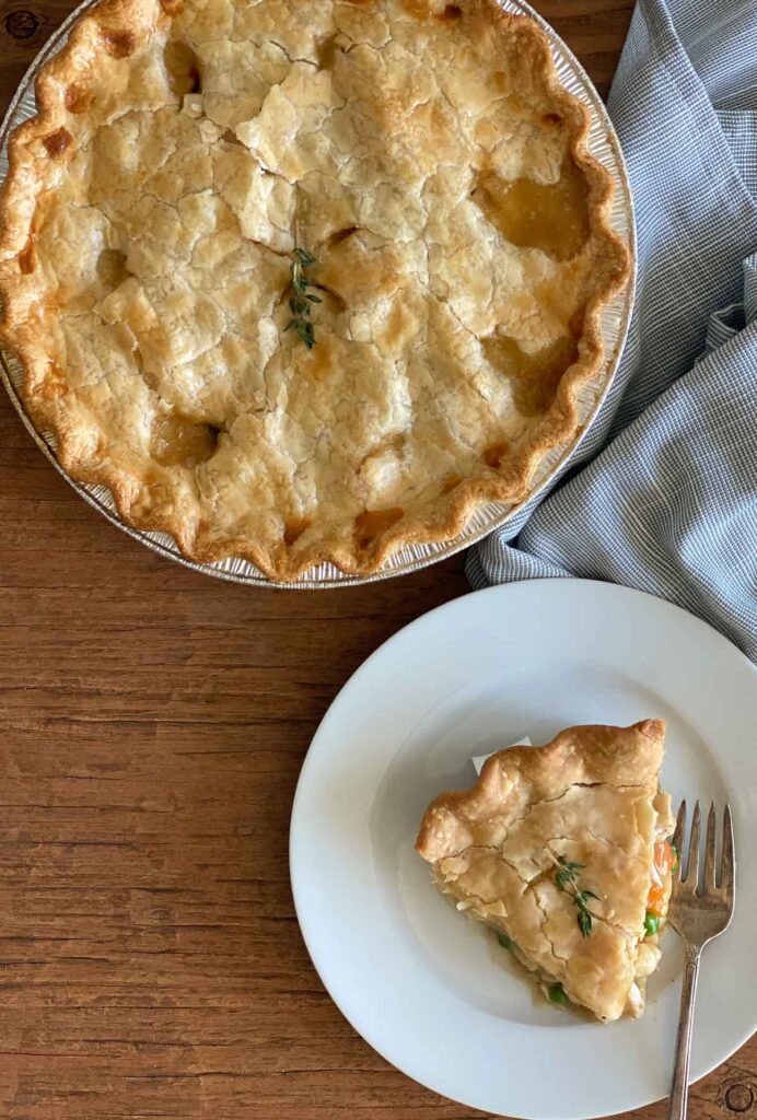On a white plate with a fork is a piece of pot pie with a whole pie in the background and a blue plaid tablecloth bunched up in the corner.