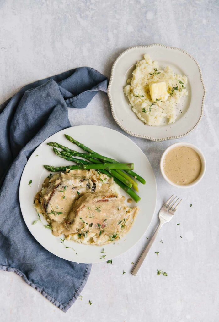 Two White plate with blue napkin on table. On plates is slow cooker pork chops covered in gravy and asparagus on the side. Other plate has mashed potatoes on it. 