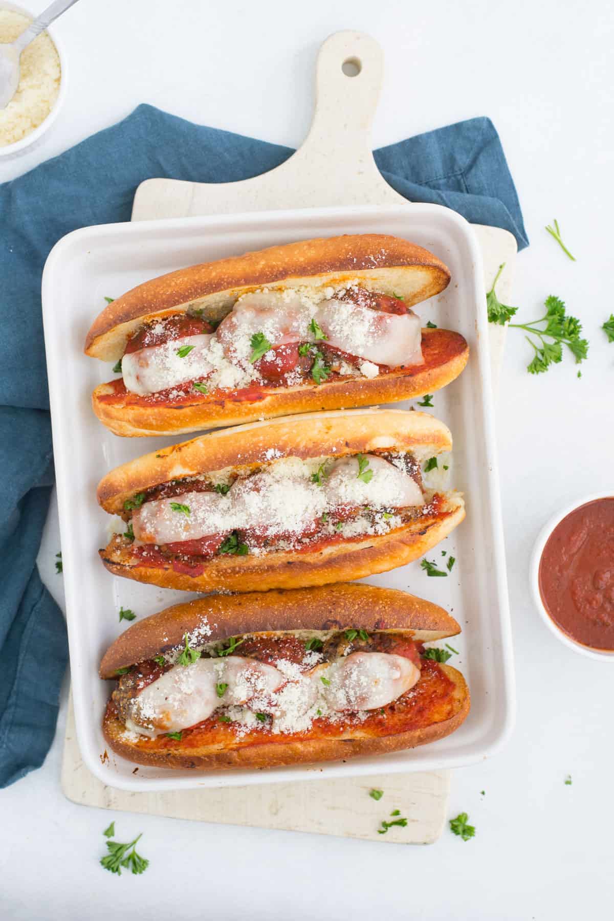 3 meatballs subs next to each other in a white dish on a white cutting board and blue napkin with a bowl of parmesan cheese in one corner and a bowl of marinara sauce in the other corner.