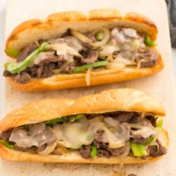 Two Philly cheesesteak sandwiches on a wooden board.
