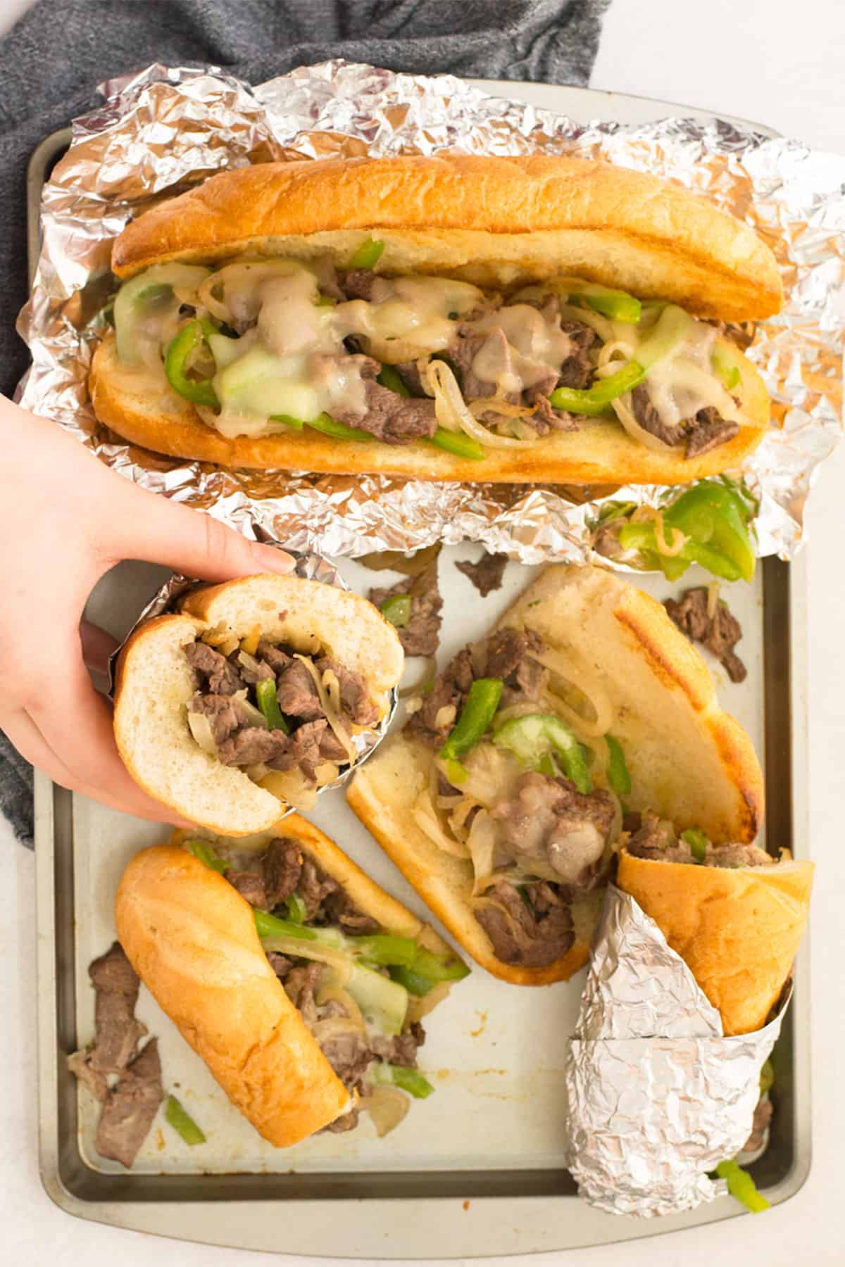 A metal pan holds a whole sandwich and four halves of a sandwich that are scattered around.  Some are wrapped in foil and one hand is holding one half of a sandwich.