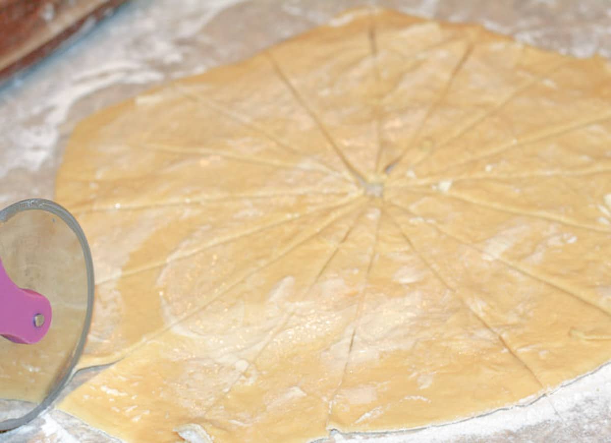 A large circular piece of rolled-out dough being cut with a pizza cutter into 16 pie shapes