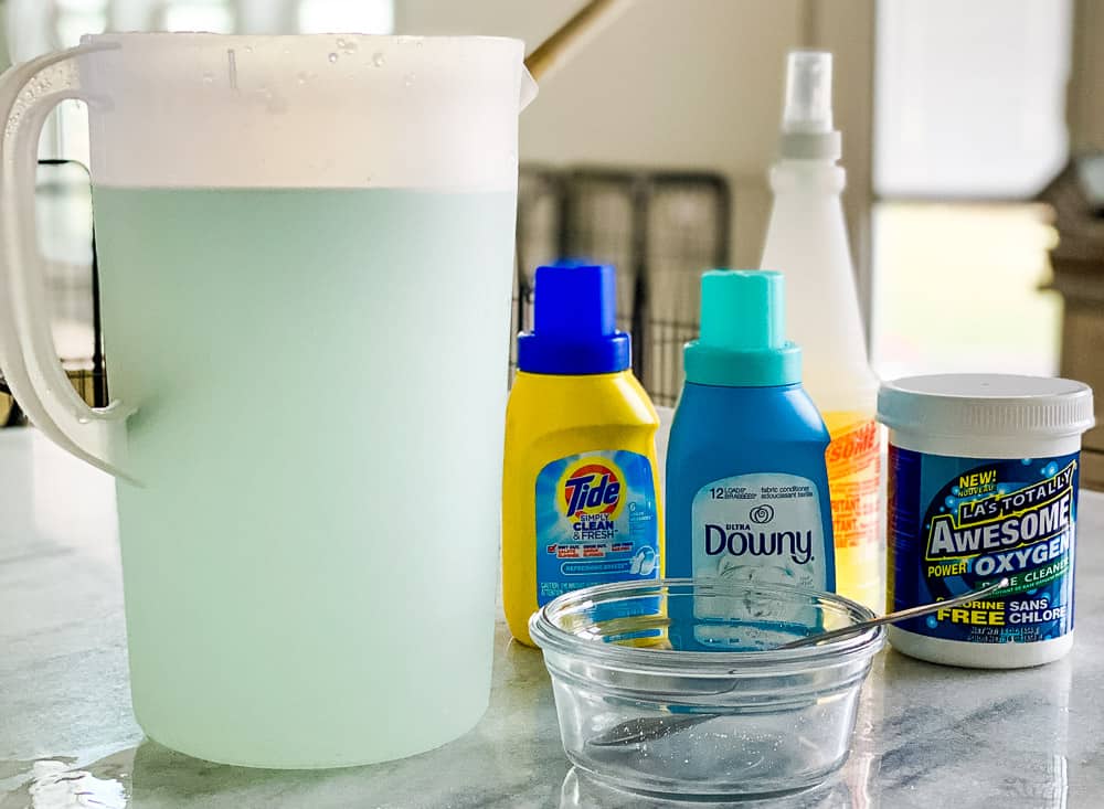 Ingredients for a recipe including oxiclean, tide, downy and awesome cleaner plus a pitcher filled with light blue solution