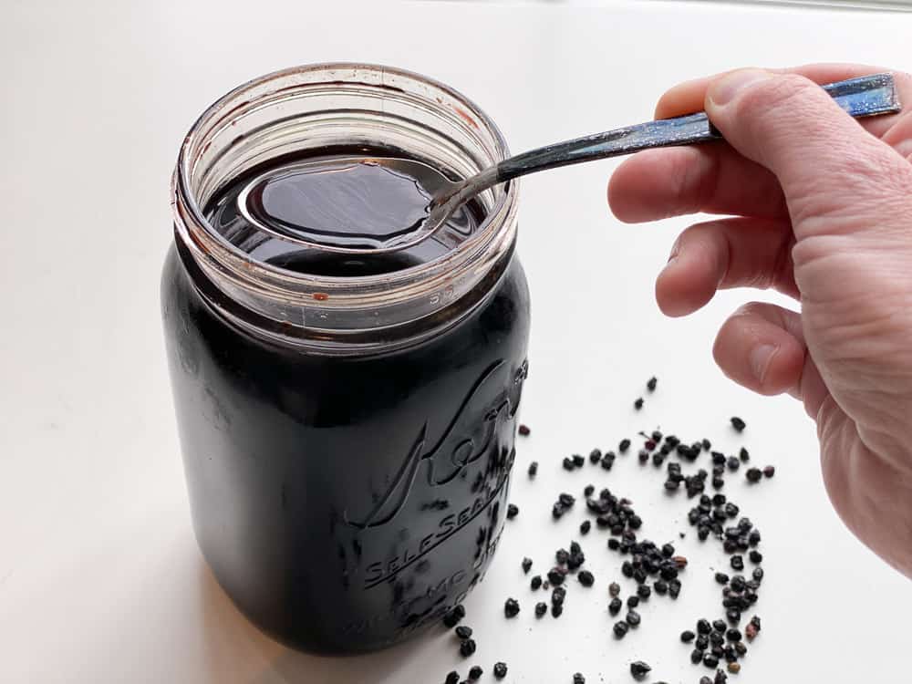 Jar of homemade elderberry syrup recipe with spoon of syrup coming out and dried elderberries beneath
