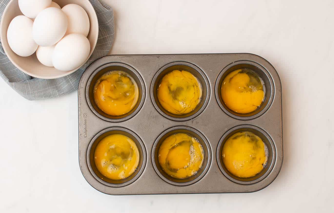 A cream bowl of whole white eggs on top of a light gray towel with a muffin tin with a single mixed-up egg in each hole.
