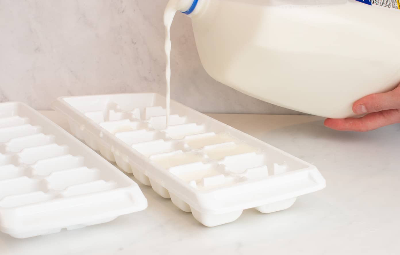 Two white ice cube trays on the white counter being filled with a gallon of milk and a hand holding it.