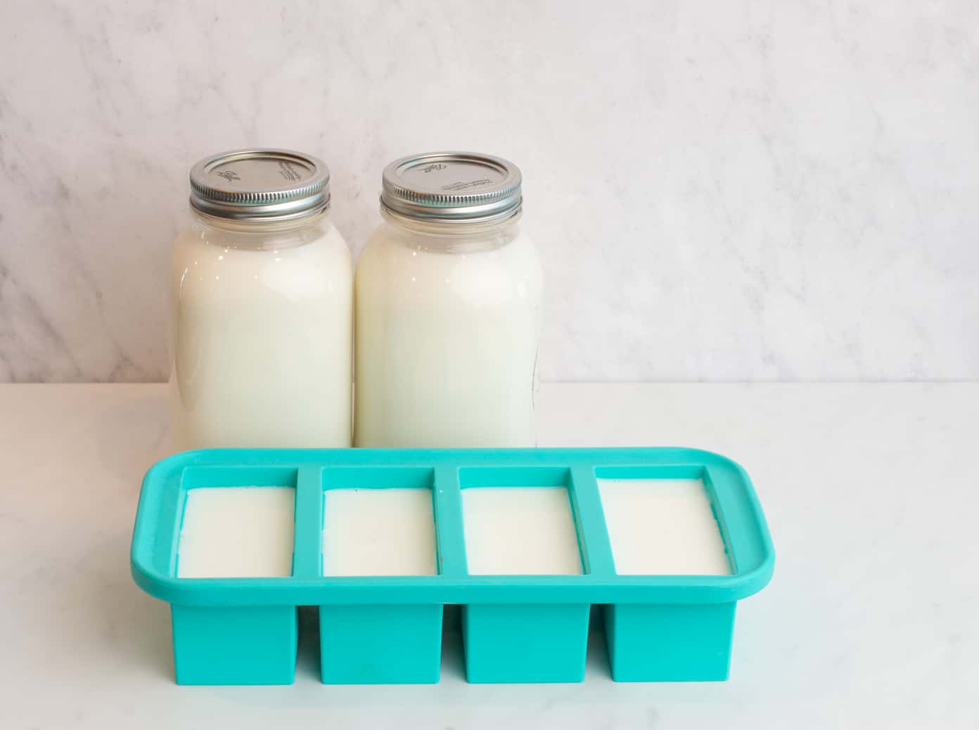 2 large mason jars lined up on a white counter and a marble background with a blue silicone container with milk in it.