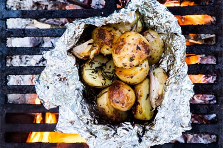A metal grate with fire underneath it has a tinfoil packet full of potatoes sprinkled with herbs. Perfect make ahead camping meals!