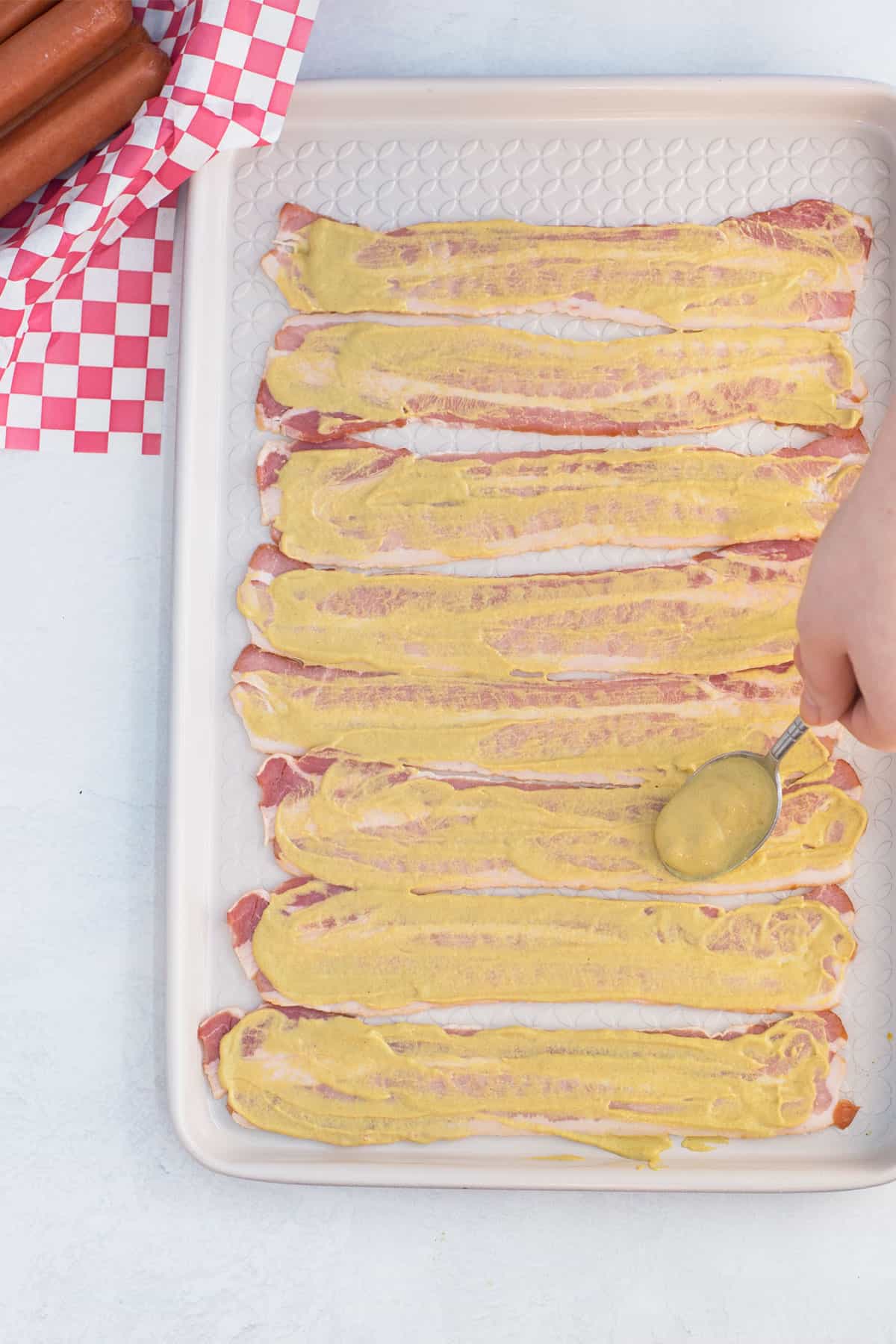 A white tray lined with bacon smothered in mustard with a hand holding a spoon of mustard spreading it on. In the corner is a red and white checked towel with raw hot dogs on it.