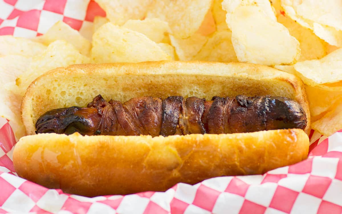 A bacon wrapped hot dog with potato chips on the side over a red and white checked paper.